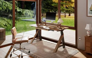 Wooden trestle desk with computer and office chair beside large windows.