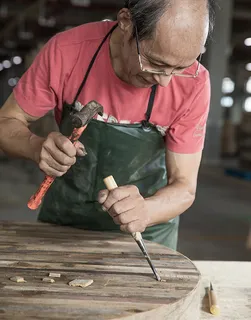Furniture worker chiseling a wooden tabletop with a hammer and chisel.